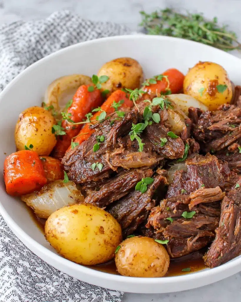Dutch oven pot roast with tender fall-apart beef chuck roast, baby carrots, baby yellow potatoes, onions, and fresh herbs in a rich savory sauce, served on a white platter.