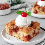 A warm slice of Dolly Parton's fruit cocktail cake with a golden crunchy top and soft fruity center, served on a white plate with a scoop of vanilla ice cream.