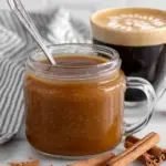 A glass jar of homemade pumpkin spice coffee syrup beside a cup of coffee on a white marble kitchen counter, with pumpkin puree, cinnamon, and warm fall spices nearby in natural window light.